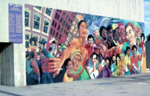 A colorful mural with diverse figures reaching outward and leaning buildings.
